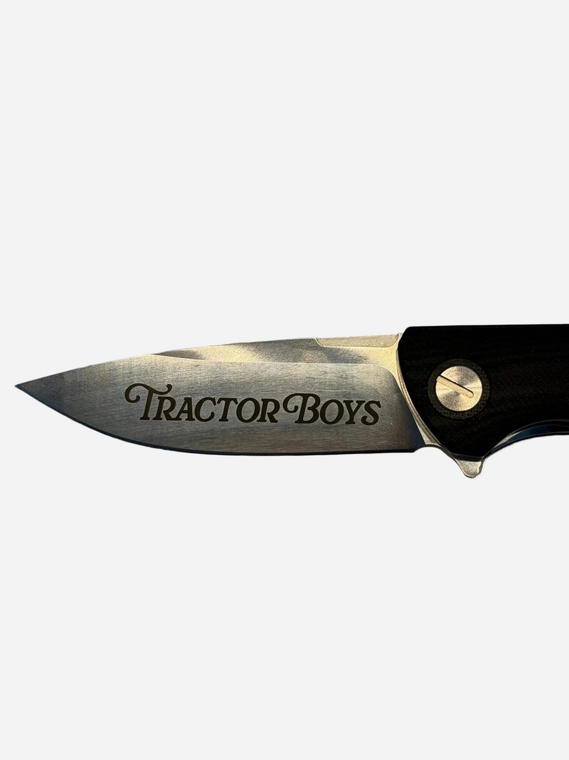 A close-up of a folding knife blade with 'TRACTOR BOYS' engraved on it, set against a light gray stone-like surface.